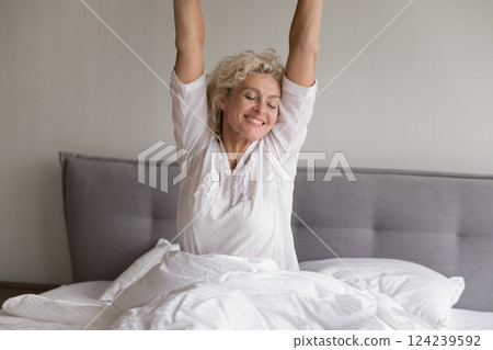 Smiling senior female sitting on bed stretching arms with pleasure 124239592