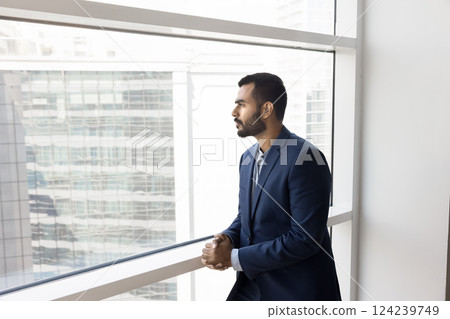 Businessman stands by office window, gazing out city skyline Businessman stands by office window, gazing out city skyline 124239749