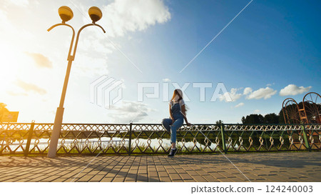 Woman perching on wooden fence, gazing at serene lake, basking in golden sunset during warm summer twilight 124240003