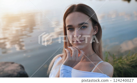 Blond woman resting hand on chin, gazing reflectively near lakeside during golden sunset lighting 124240027