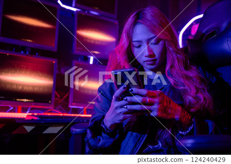 Asian woman in front of a computer monitor display using mobile phone, neon light, Hacker wear black jacket at system control room, Security guard working on multiple monitors together smartphone 124240429