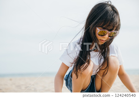 Beach style fashion, a child girl with trendy sunglasses sitting on a deck chair. Smiling portrait radiates joy and happiness. Windy hair stylish beachwear and carefree relaxation 124240540