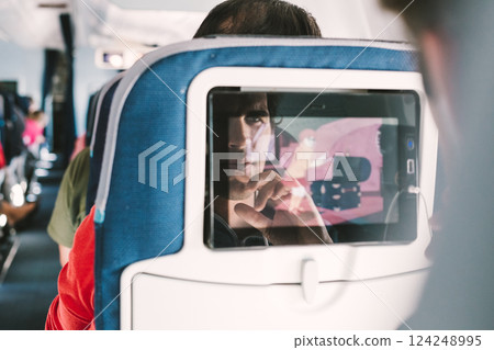 Reflection of a passenger on an airplane touch screen monitor while watching cartoon during long flight. Entertainment service system in aircraft Reflection of a passenger on an airplane touch screen monitor while watching cartoon during long flight. Entertainment service system in aircraft 124248995