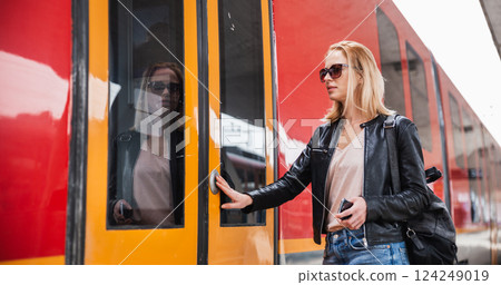 Young blond woman in jeans, shirt and leather jacket wearing bag and sunglass, presses door button of modern speed train to board on train station platform. Travel and transportation. 124249019