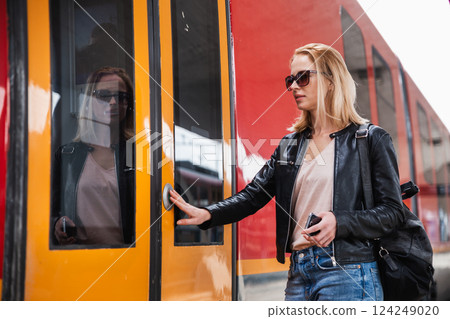 Young blond woman in jeans, shirt and leather jacket wearing bag and sunglass, presses door button of modern speed train to board on train station platform. Travel and transportation. 124249020