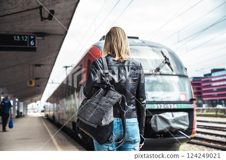 Young blond woman in jeans, shirt and leather jacket wearing bag and sunglass, embarking red modern speed train on train station platform. Travel and transportation 124249021