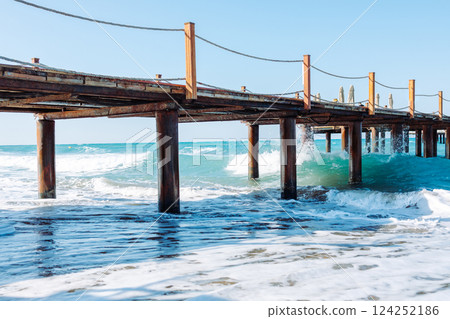 Picturesque seascape with a wooden pier extending into the turquoise waters of the Mediterranean Sea. Powerful waves crash against rusty metal supports. Azure sky. Picturesque seascape with a wooden pier extending into the turquoise waters of the Mediterranean Sea. Powerful waves crash against rusty metal supports. Azure sky. 124252186