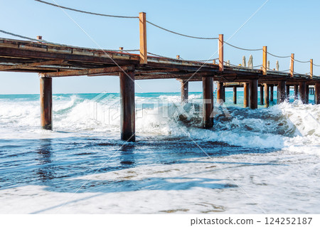 Picturesque seascape with a wooden pier extending into the turquoise waters of the Mediterranean Sea. Powerful waves crash against rusty metal supports. Azure sky. Picturesque seascape with a wooden pier extending into the turquoise waters of the Mediterranean Sea. Powerful waves crash against rusty metal supports. Azure sky. 124252187