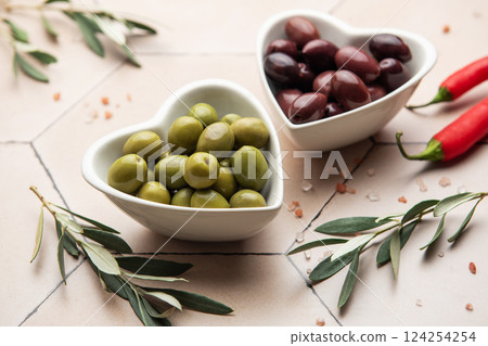 Heart shaped bowls containing green and black olives, olive branches, chili peppers and himalayan salt on tiled surface 124254254