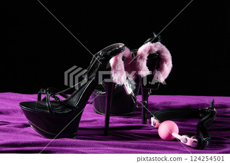 Adult sex games. Kinky lifestyle. Pink gag ball, handcuffs and a pair of black high-heeled shoes on the purple bedsheet. Bdsm outfit Adult sex games. Kinky lifestyle. Pink gag ball, handcuffs and a pair of black high-heeled shoes on the purple bedsheet. Bdsm outfit 124254501