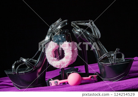 Adult sex games. Kinky lifestyle. Pink gag ball, handcuffs and a pair of black high-heeled shoes on the purple bedsheet. Bdsm outfit Adult sex games. Kinky lifestyle. Pink gag ball, handcuffs and a pair of black high-heeled shoes on the purple bedsheet. Bdsm outfit 124254529