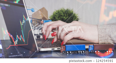 Dynamic E-Commerce and Stock Market Analysis Workspace Featuring Hands on Laptop with Candlestick Chart, Miniature Toy Trucks, Shopping Cart, and Green Plant in Modern Office Setting 124254591