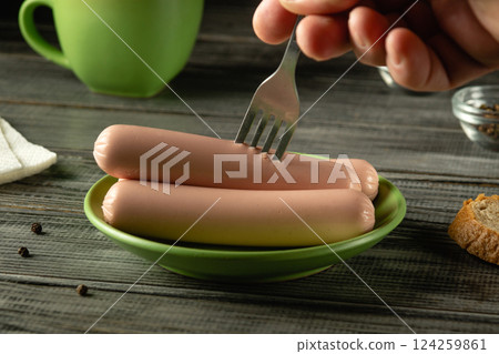 A hand holds a fork to pierce cooked sausages resting on a green plate placed on a rustic wooden table. Surrounding items include a green mug and a slice of bread A hand holds a fork to pierce cooked sausages resting on a green plate placed on a rustic wooden table. Surrounding items include a green mug and a slice of bread 124259861