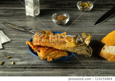 Golden crispy fried fish rests on a blue plate alongside slices of rustic bread. Condiments are placed on a wooden table, creating an appetizing display 124259864