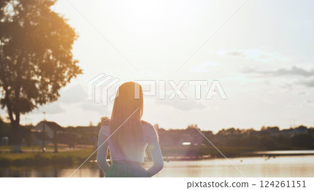 Blonde schoolgirl contemplating golden sunset hues reflecting across calm lake waters, framed by residential neighborhood homes 124261151
