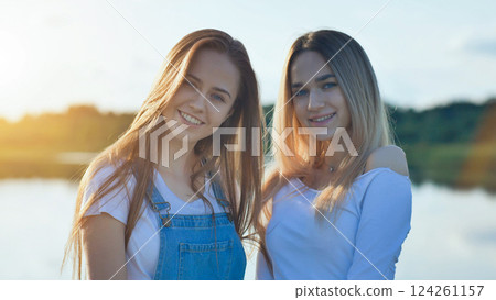 Teenage friends smiling together near tranquil lakeside during golden sunset, experiencing joyful summer friendship moment 124261157