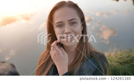 Young student resting near lakeside, wearing denim jacket, softly touching cheek while smiling during golden sunset light 124261160
