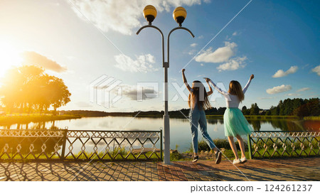 Teenage female friends leaping near lakeside during golden hour, marking school year completion and summer break anticipation Teenage female friends leaping near lakeside during golden hour, marking school year completion and summer break anticipation 124261237