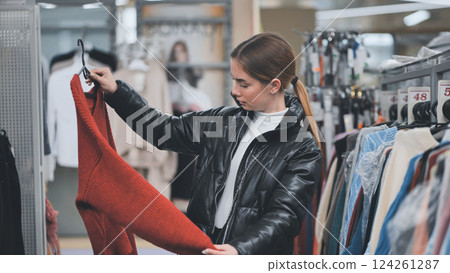 Young woman browsing clothing store, selecting red sweater on hanger while examining trendy fashion displays Young woman browsing clothing store, selecting red sweater on hanger while examining trendy fashion displays 124261287