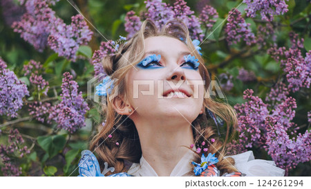 Female model sporting butterfly lashes, vibrant makeup, standing amid blooming lilac garden, inhaling springtime floral sweetness 124261294