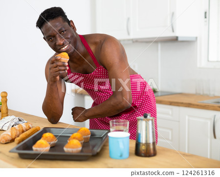 Cheerful naked man in red apron tasting muffin in home kitchen Cheerful naked man in red apron tasting muffin in home kitchen 124261316