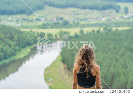 Rear view of a hiker woman admiring a serene river and lush green valley while standing atop a breathtaking mountain 124261400