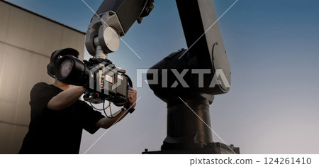 Video or film production with high tech robotic arm equipped with a professional cinema camera. Operated by a cinematographer. Advanced motion control system enables precise camera movements filming. 124261410