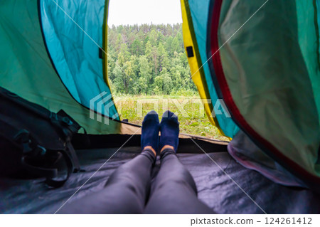Camper relaxing in tent, enjoying serene woodland vista with sunlight filtering through verdant trees 124261412