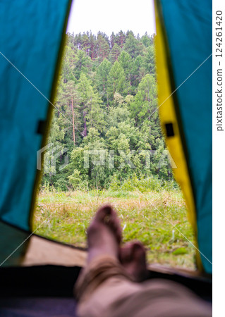Legs of a camper relaxing inside a tent, soaking in the stunning view of a lush green forest and enjoying a moment of tranquility Legs of a camper relaxing inside a tent, soaking in the stunning view of a lush green forest and enjoying a moment of tranquility 124261420