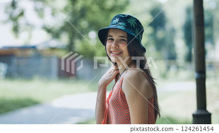 Cheerful teen with bright smile wearing bucket hat, basking in summer sunshine amid verdant park landscape Cheerful teen with bright smile wearing bucket hat, basking in summer sunshine amid verdant park landscape 124261454