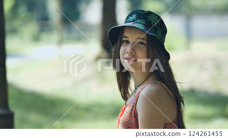 Teenage girl in marijuana leaf bucket hat relaxing outdoors, natural summer park lighting highlighting casual youth style Teenage girl in marijuana leaf bucket hat relaxing outdoors, natural summer park lighting highlighting casual youth style 124261455