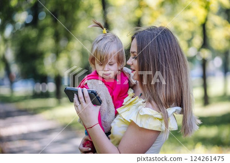 Mother holding her daughter and showing her a smartphone in a park, enjoying time together 124261475