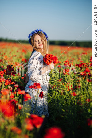 Golden haired woman wearing floral headpiece, carrying red poppy bouquet amid blooming poppy field during sunset light 124261487