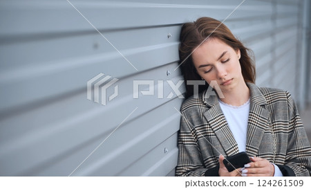 Dejected professional resting against metallic wall, staring intently at smartphone, embodying workplace isolation and emotional strain 124261509