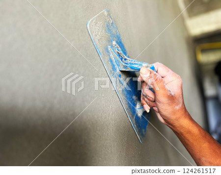 Construction worker plastering a wall with a blue trowel. The image captures the process of smoothing cement for a perfect finish in building construction Construction worker plastering a wall with a blue trowel. The image captures the process of smoothing cement for a perfect finish in building construction 124261517