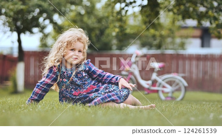Blond child lounging on lawn, bicycle softly blurred, basking in warm sunlight during carefree family afternoon Blond child lounging on lawn, bicycle softly blurred, basking in warm sunlight during carefree family afternoon 124261539