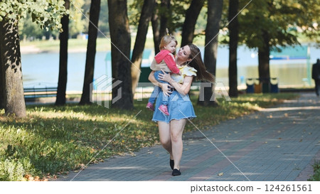 Mother cuddling young daughter while strolling lakeside park during bright summer afternoon 124261561