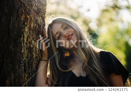 Blonde teenage girl leaning against tree trunk, smiling while sunlight filters through green leaves during warm summer day 124261571