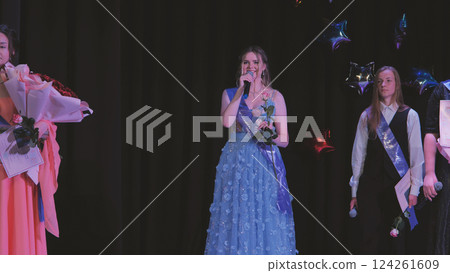 High school graduate delivering commencement speech, clutching rose and microphone, surrounded by classmates in graduation attire 124261609