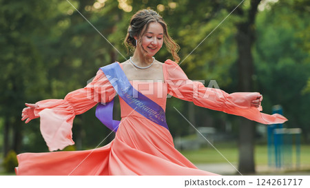 Graduate student wearing a purple sash twirling in a coral dress, celebrating graduation in a park Graduate student wearing a purple sash twirling in a coral dress, celebrating graduation in a park 124261717