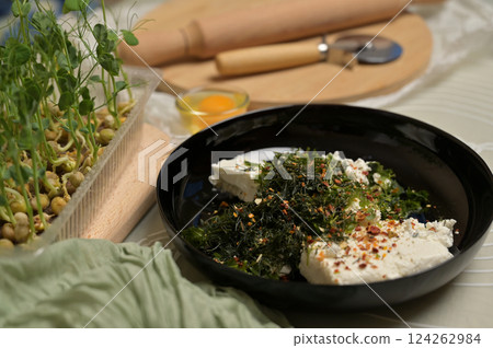 Parsley and dill greens with cottage cheese in a black bowl. Rolling pin, cutter, eggs, green towel and microgreens sprouts on kitchen table. Home cooking baking puffs 124262984