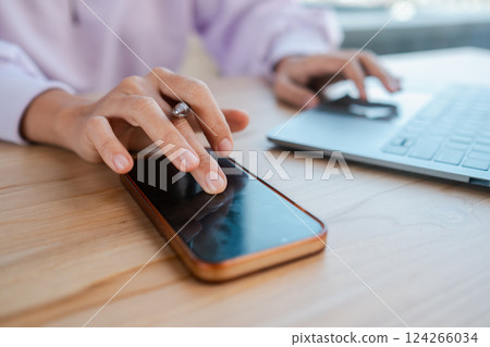 Hands interacting with smartphone and laptop on a wooden desk, showcasing multitasking in a modern workspace. Hands interacting with smartphone and laptop on a wooden desk, showcasing multitasking in a modern workspace. 124266034