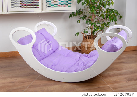 White wooden rocking bed adorned with violet cushions in a cozy indoor setting. Kids rocking bed placed in a warmly lit corner of a room with natural flooring and indoor decor 124267037