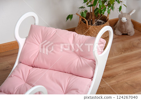 White wooden rocking bed adorned with pink cushions in a cozy indoor setting. Kids rocking bed placed in a warmly lit corner of a room with natural flooring and indoor decor White wooden rocking bed adorned with pink cushions in a cozy indoor setting. Kids rocking bed placed in a warmly lit corner of a room with natural flooring and indoor decor 124267040