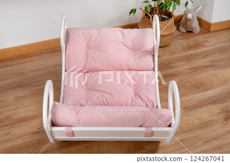 White wooden rocking bed adorned with pink cushions in a cozy indoor setting. Kids rocking bed placed in a warmly lit corner of a room with natural flooring and indoor decor White wooden rocking bed adorned with pink cushions in a cozy indoor setting. Kids rocking bed placed in a warmly lit corner of a room with natural flooring and indoor decor 124267041