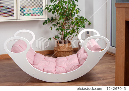 White wooden rocking bed adorned with pink cushions in a cozy indoor setting. Kids rocking bed placed in a warmly lit corner of a room with natural flooring and indoor decor White wooden rocking bed adorned with pink cushions in a cozy indoor setting. Kids rocking bed placed in a warmly lit corner of a room with natural flooring and indoor decor 124267043