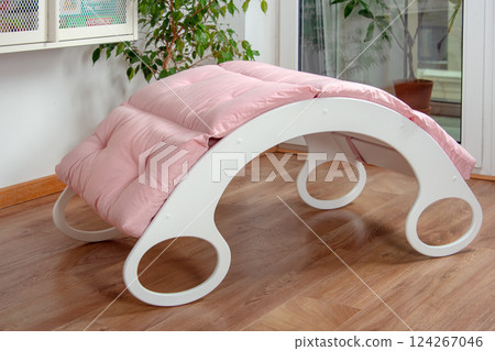White wooden rocking bed adorned with pink cushions in a cozy indoor setting. Kids rocking bed placed in a warmly lit corner of a room with natural flooring and indoor decor 124267046