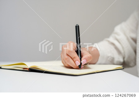 a person's hand writing with a black pen in an open notebook on a white desk a person's hand writing with a black pen in an open notebook on a white desk 124267069