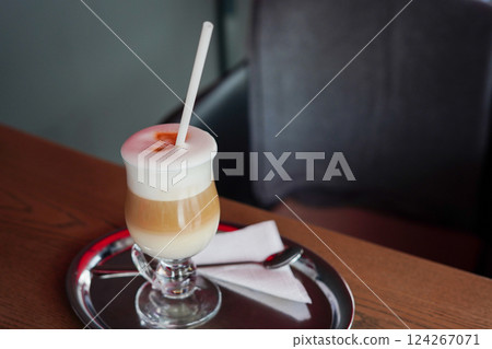 latte macchiato served in a clear glass with a straw, placed on a silver tray with a napkin and spoon on a wooden table 124267071