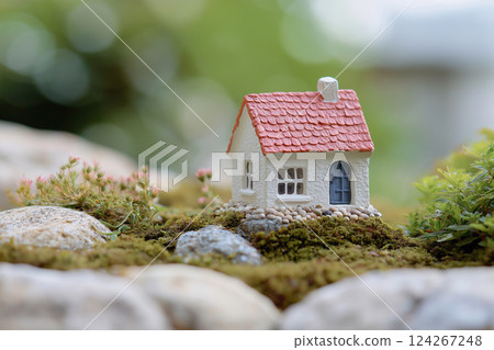 A miniature model of a house with a red roof is placed on a bed of gray stones, surrounded by moss and small green plants, set against a softly blurred natural background. 124267248